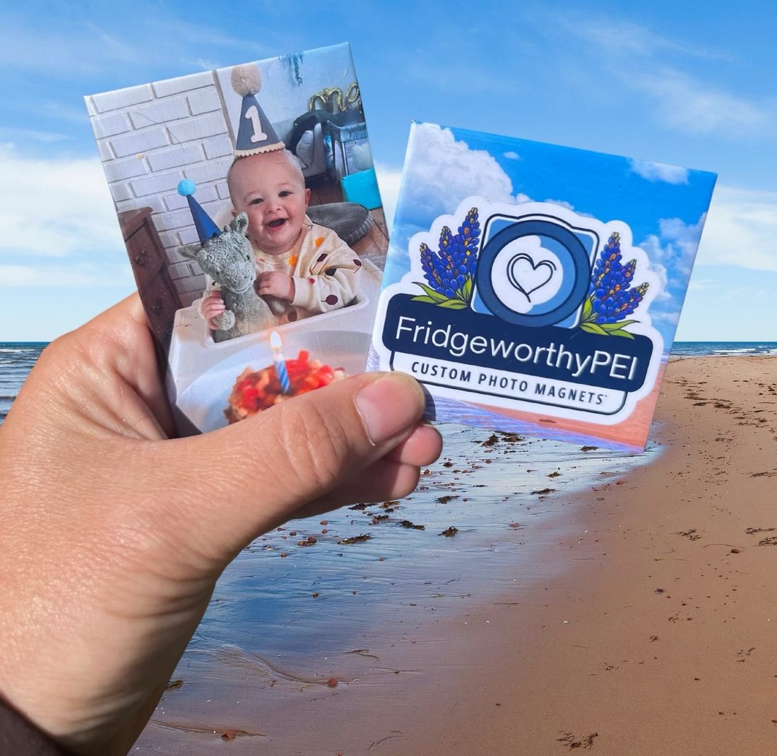 A hand holding two Fridgeworthy photo magnets on a PEI red sand beach
